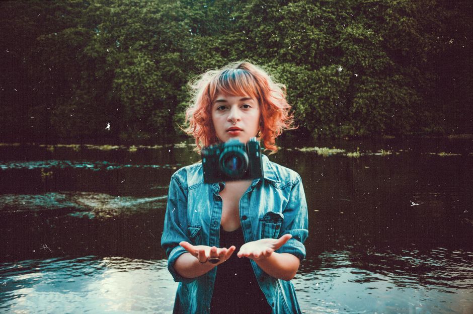 A young woman with red hair and denim jacket throwing a camera near a river.