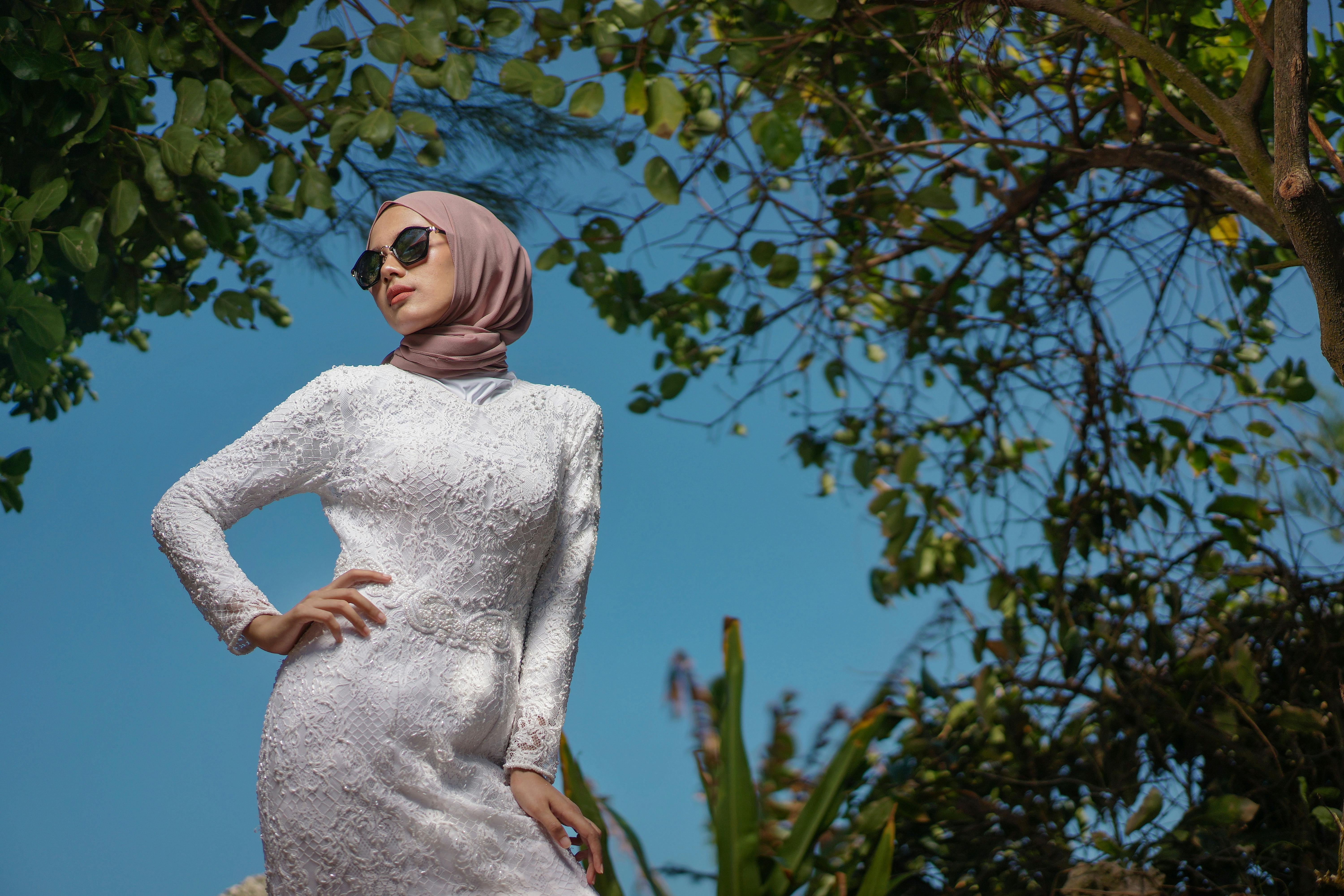 Stylish woman in sunglasses and hijab posing outdoors against a leafy backdrop.