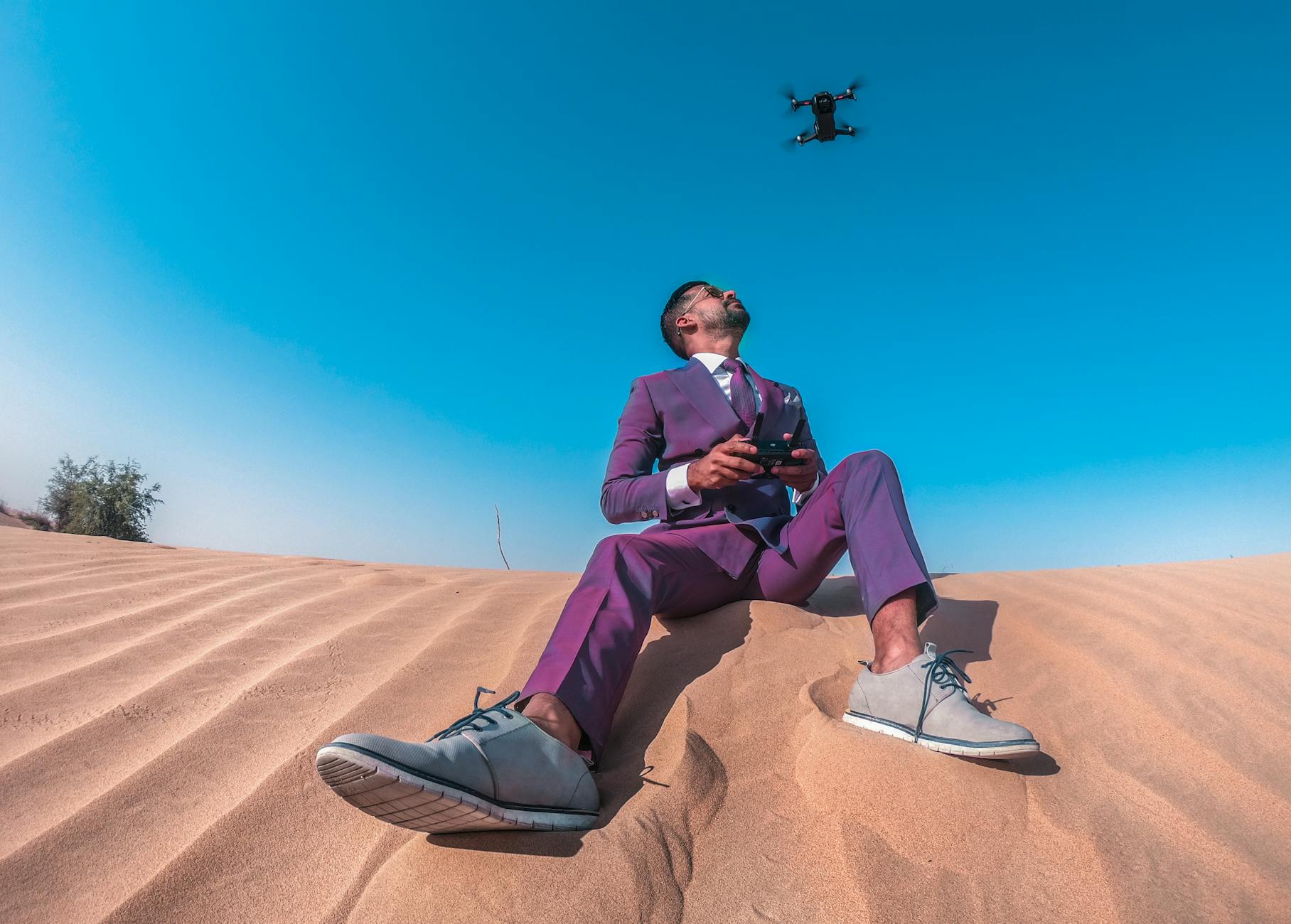 A stylish man in a suit controls a drone against a clear blue sky in the Dubai desert.