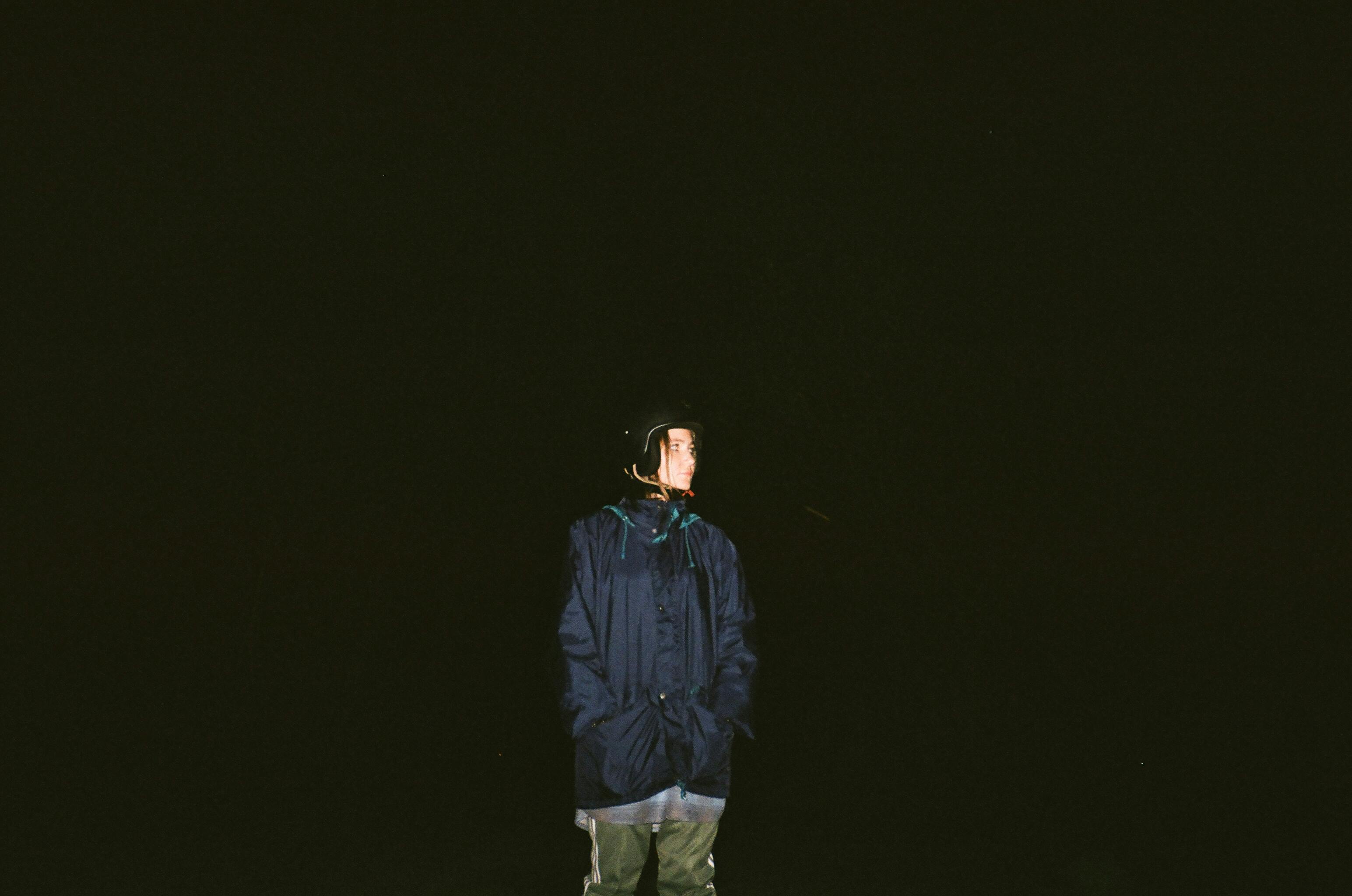 一个孤独的身影穿着深色夹克，站在漆黑的背景中，在夜晚的灯光下被照亮