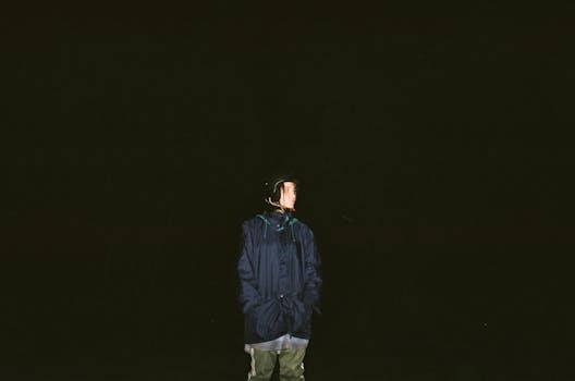 一个孤独的身影穿着深色夹克，站在漆黑的背景中，在夜晚的灯光下被照亮