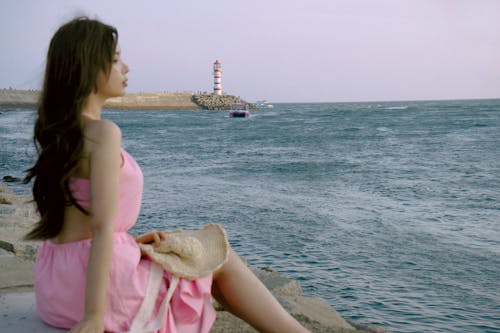 A woman in a pink dress sitting on a rock by the water