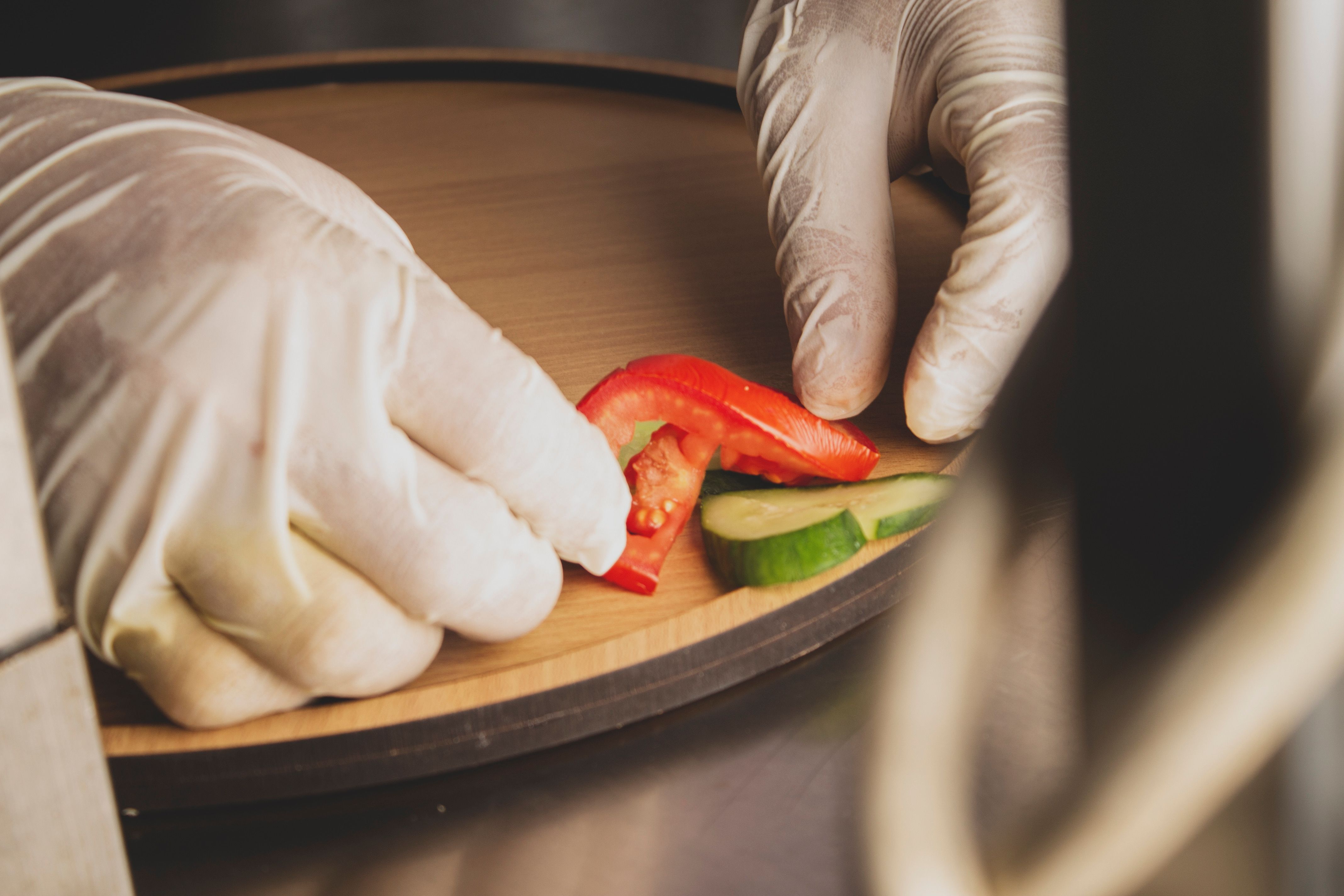 Close-up of gloved hands arranging cucumber and tomato slices on a wooden tray.