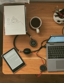 A minimalistic workspace featuring a laptop, e-reader, headphones, and coffee on a wooden desk.