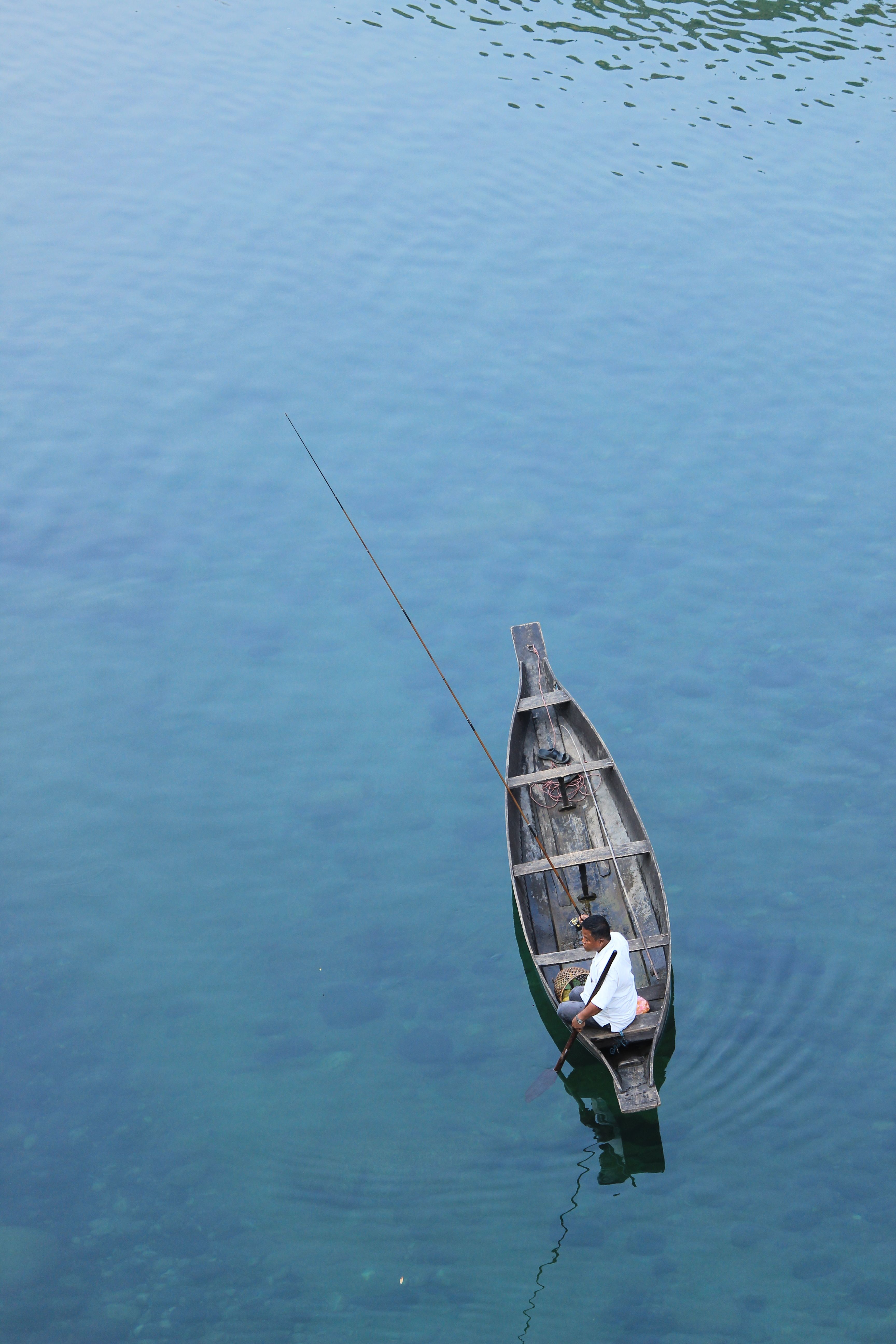 A man fishing from a wooden boat on a crystal-clear lake, captured from above.