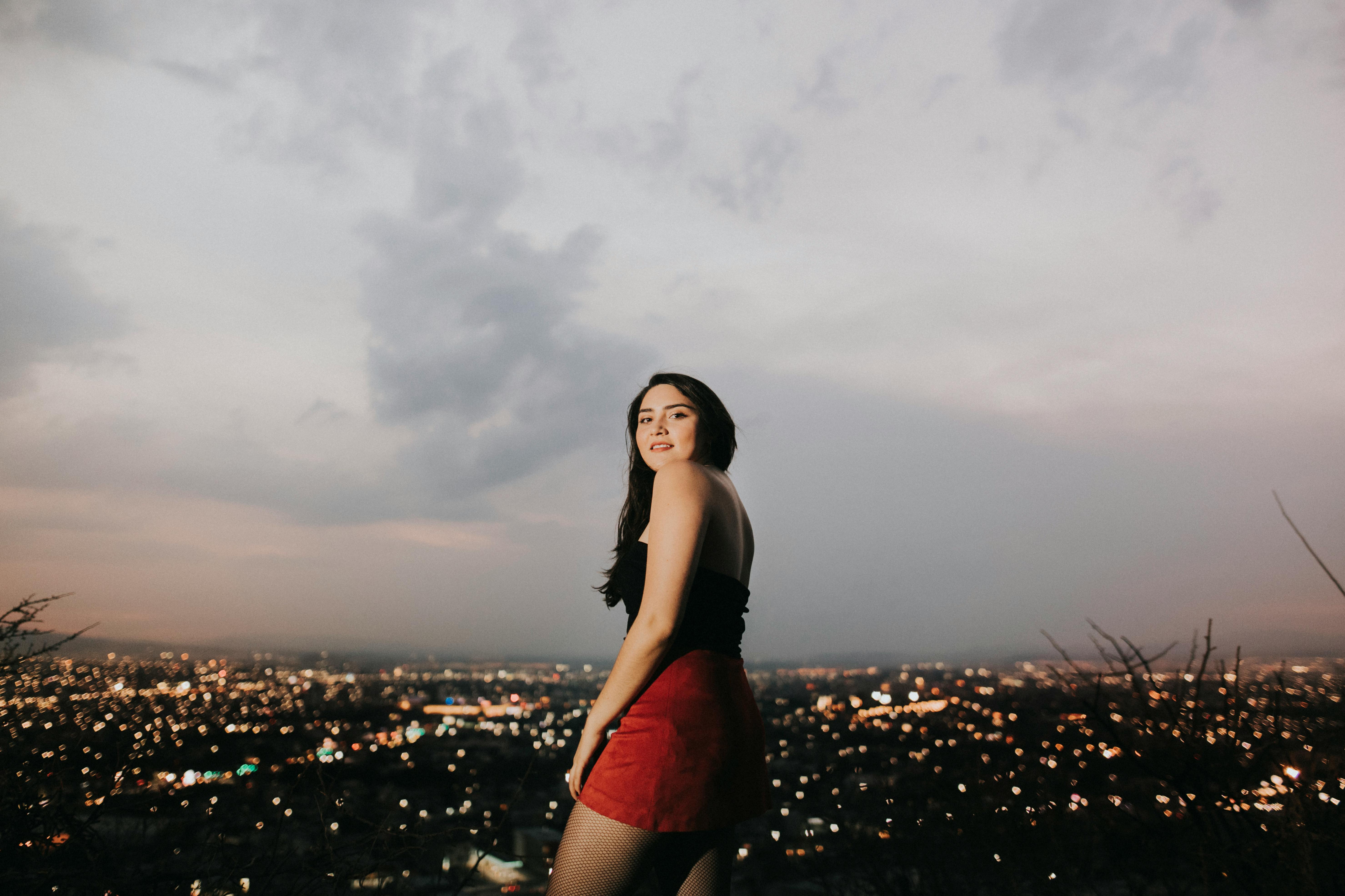 黄昏时分，一名年轻女子站在山上，俯瞰着城市灯火与宁静夜空融为一体的景象