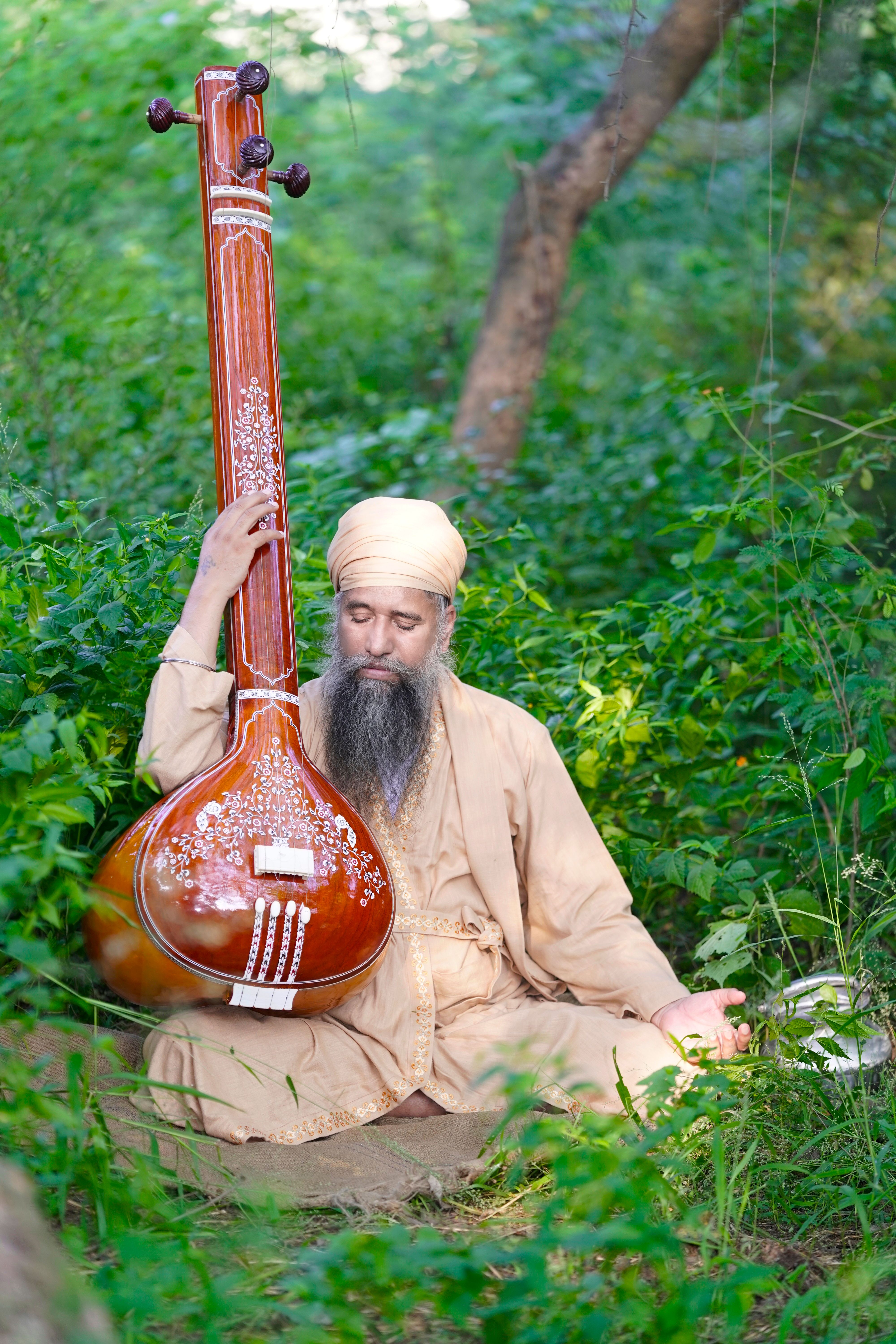 A bearded man in traditional attire plays tanpura amidst lush greenery in an Indian forest.