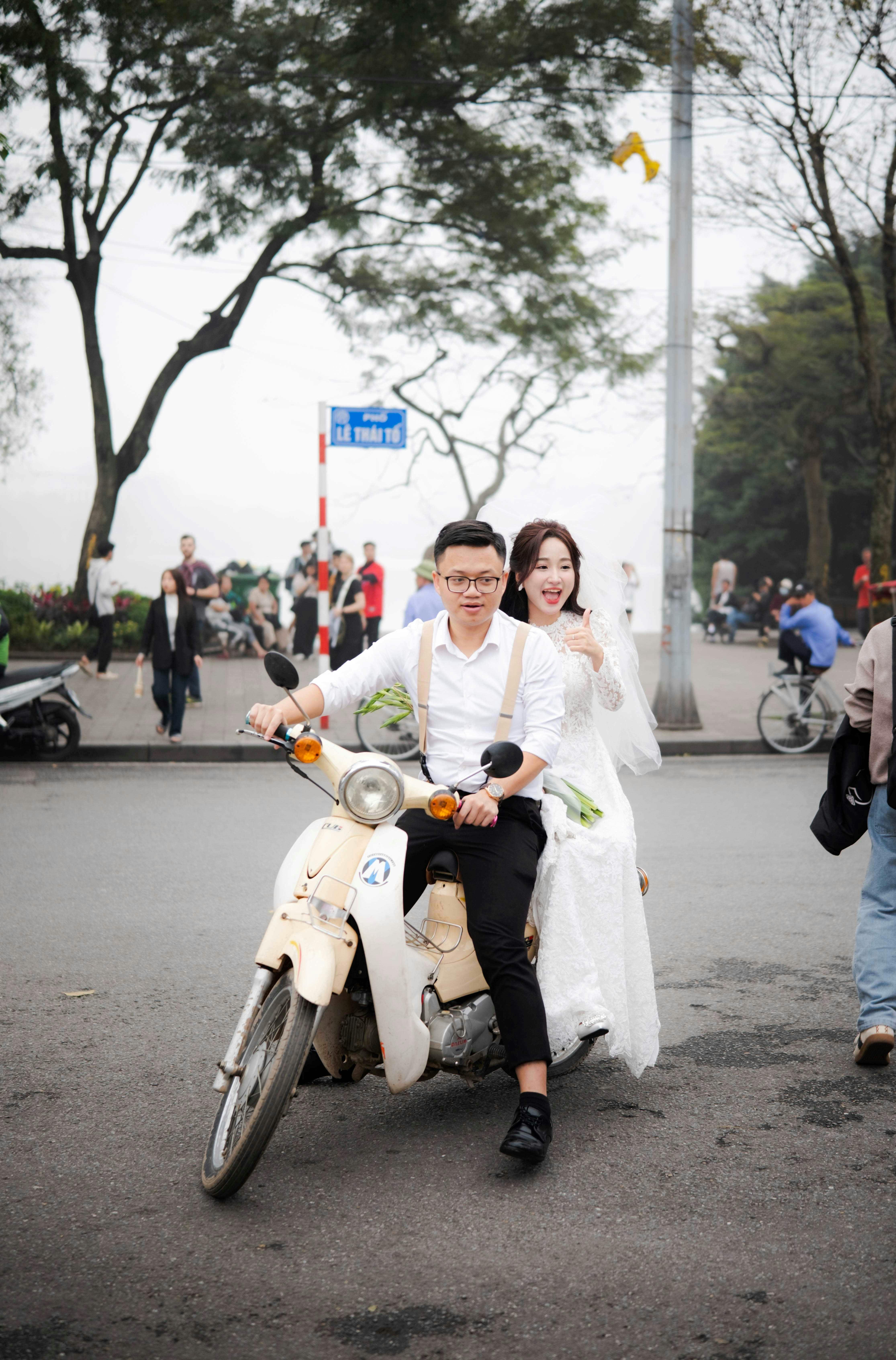 Smiling couple in wedding attire riding a scooter through busy urban streets.