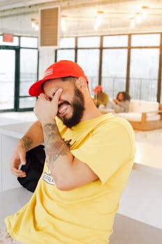 一个快乐的男子头戴红帽、身穿黄衬衫，在室内开怀大笑，散发着幸福与放松的气息