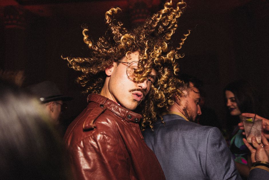 Energetic man with curly hair flips head in lively nightclub setting, expressing fun.