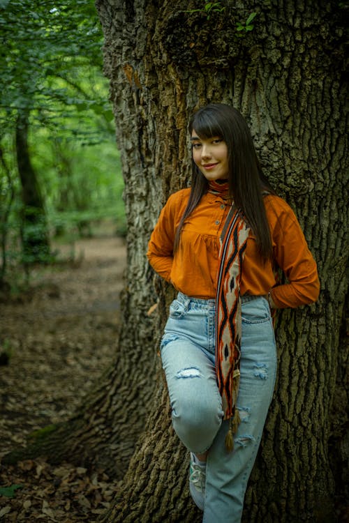 A girl in an orange shirt and jeans leaning against a tree