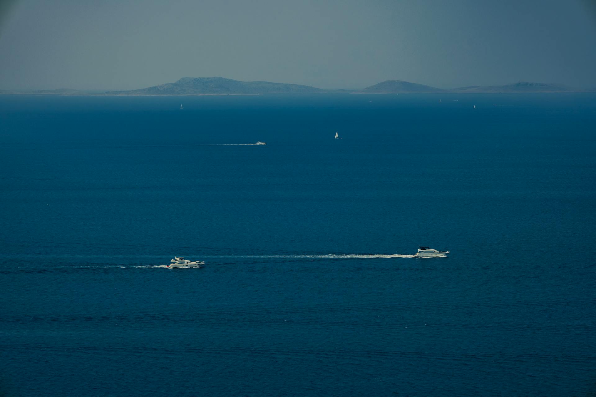 Aerial view of yachts on the deep blue ocean with distant islands under a clear sky.