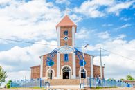 A picturesque Catholic church with a distinctive red roof and blue accents under a vibrant sky.