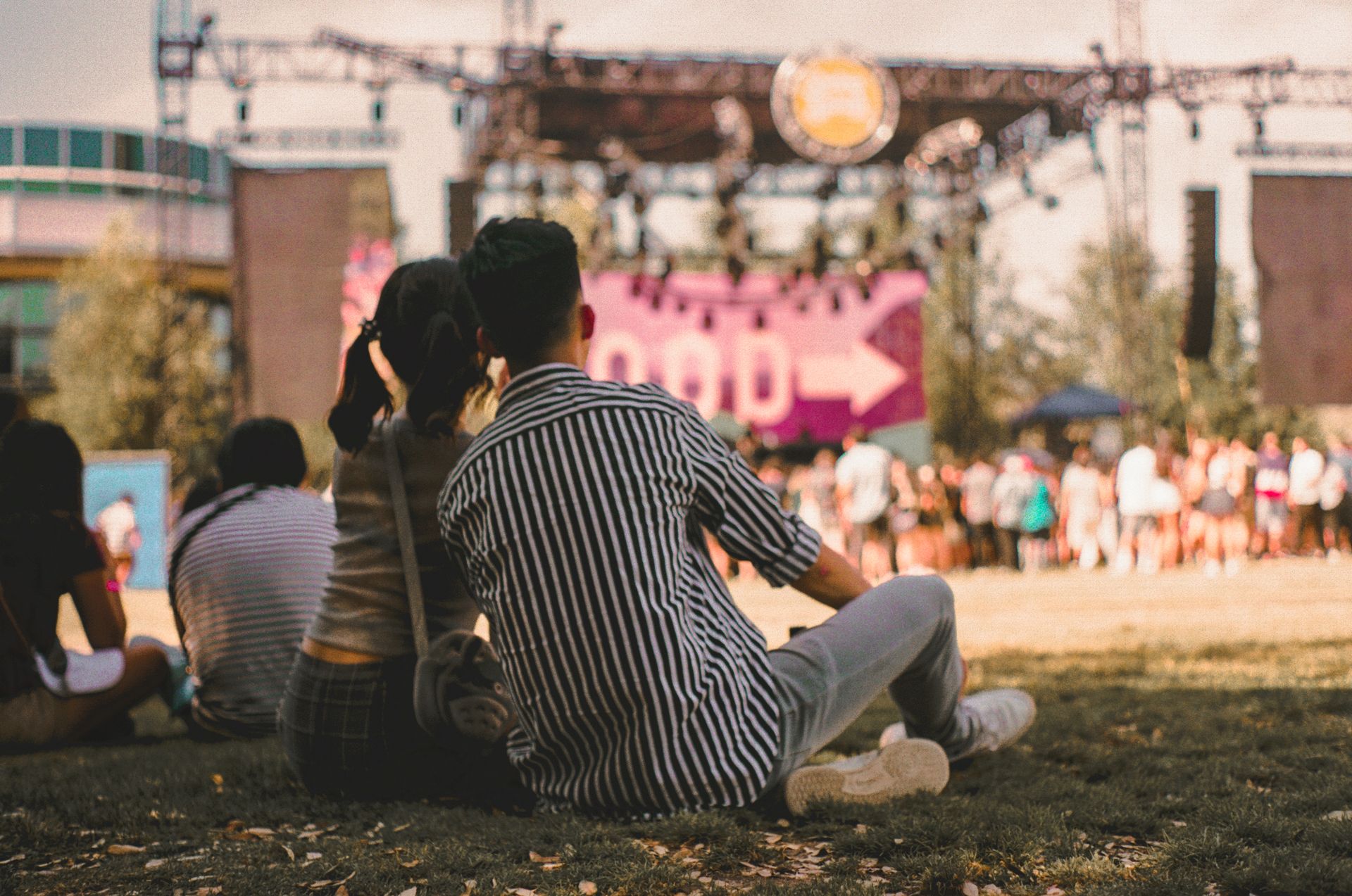 A couple sits on the grass, enjoying a sunny day at a lively outdoor music festival.