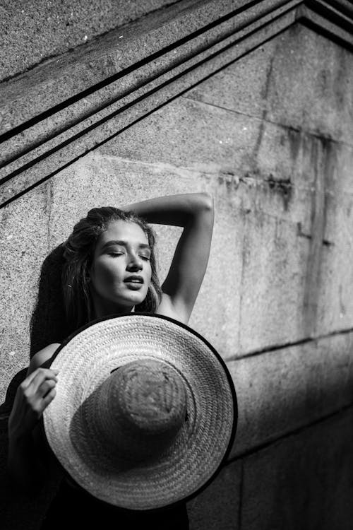 Monochrome Photo of Woman Leaning on Wall While Holding Sun Hat
