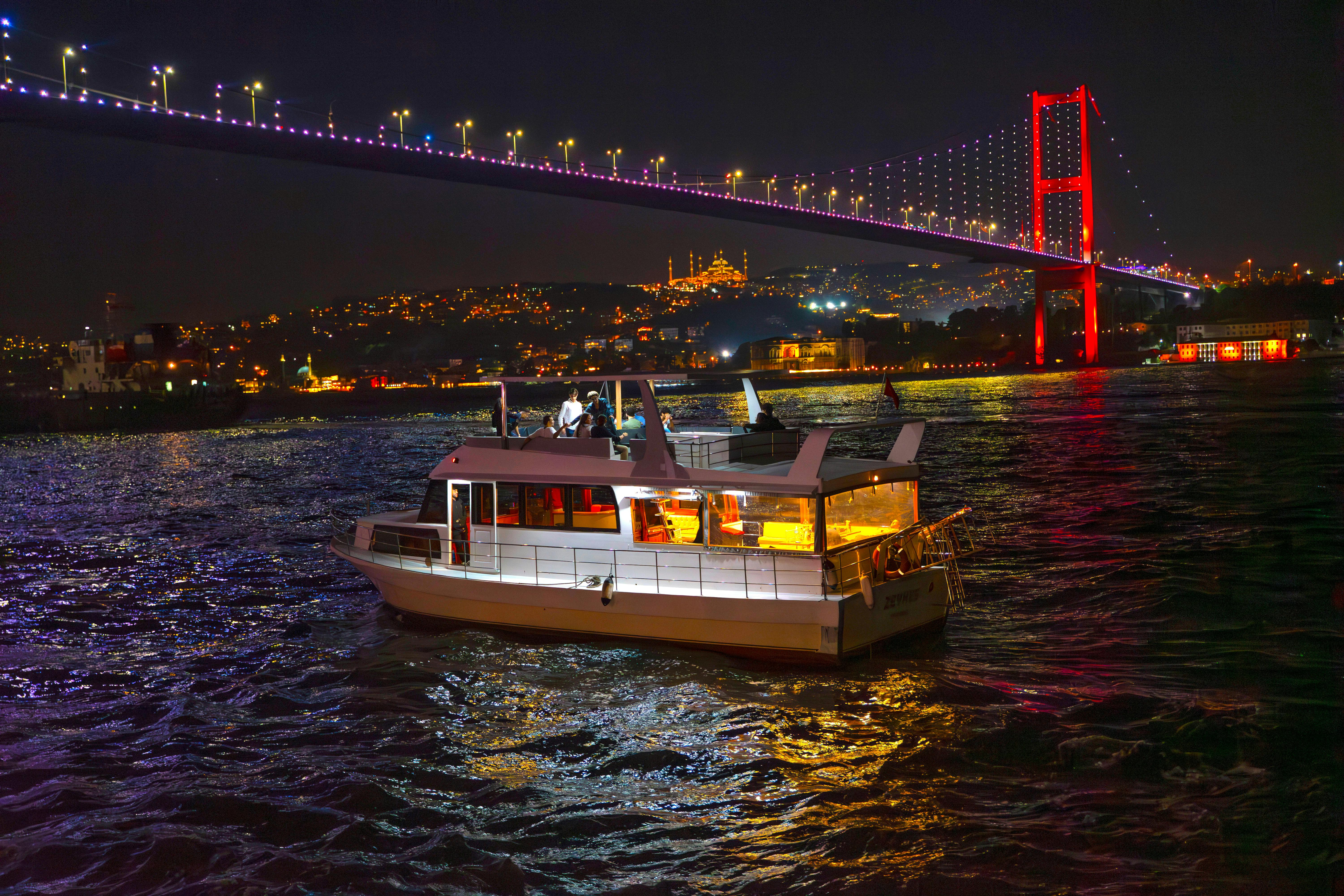 A motor yacht cruises under Istanbul's illuminated Bosphorus Bridge at night, showcasing the vibrant