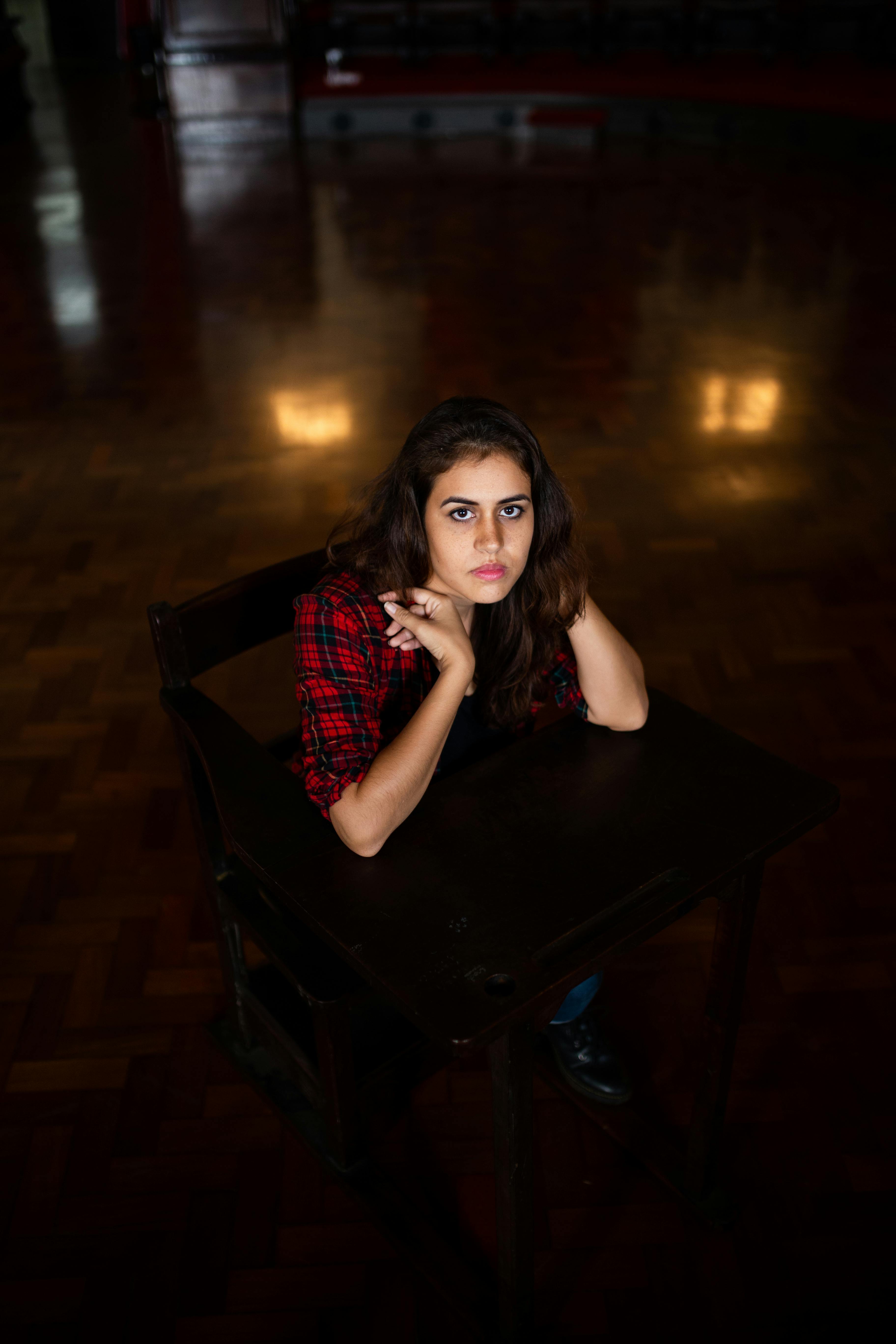 一位穿着格子衬衫的年轻女子坐在光线昏暗的房间的木桌前，脸上流露出若有所思的表情