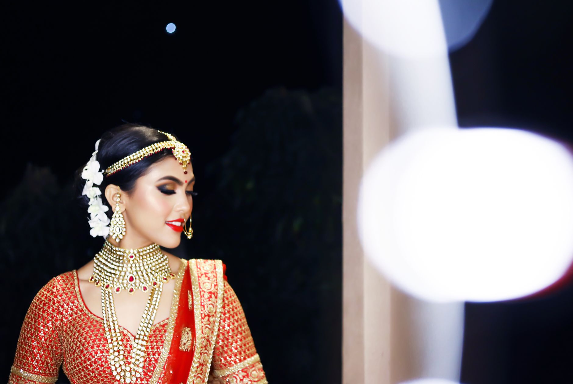 Indian bride in traditional red and gold attire at night with elaborate jewelry and makeup.