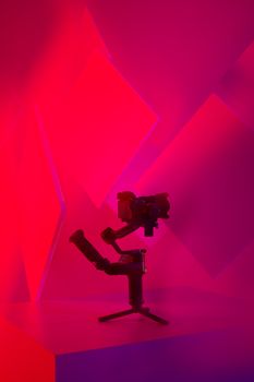A sleek camera gimbal displayed in a studio with striking red and pink lighting.