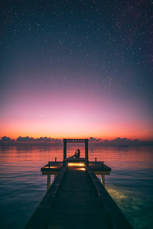 Silhouette Photo of Person Sitting on Boardwalk At Sunset