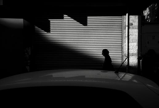 Black and white street photography capturing silhouette of a person walking past urban shadows.