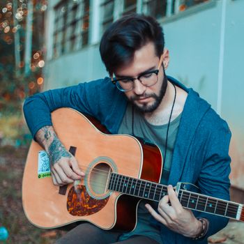 A young man with tattoos playing an acoustic guitar in a serene outdoor setting.