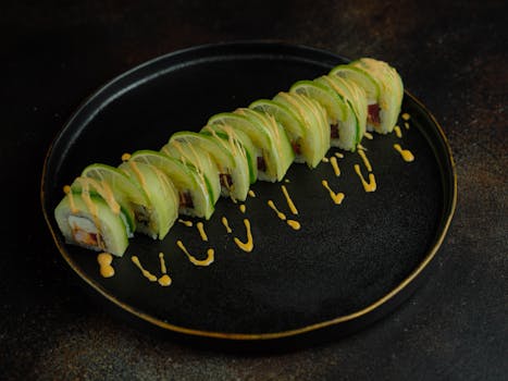 Close-up of gourmet sushi rolls topped with avocado and lime, drizzled with sauce on a black plate.