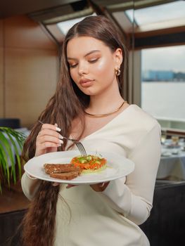 A young brunette woman in a white dress savoring a gourmet dish by a window indoors.