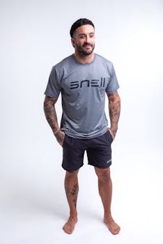 Portrait of a tattooed man in a gray t-shirt and shorts, standing barefoot against a white background.
