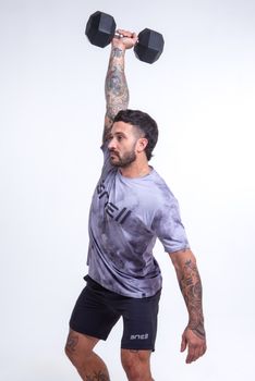 Tattooed man lifting dumbbell in a studio, sporting gray sportswear on white background.