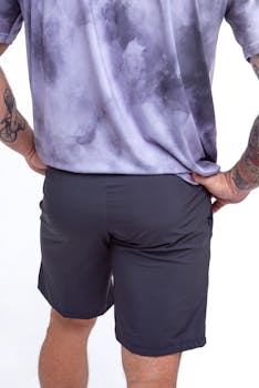 A man poses in a purple t-shirt and shorts against a white background, showcasing fashion and tattoos.