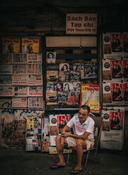 Free A man sits by a newspaper and magazine stall on an urban street, displaying diverse publications. Stock Photo