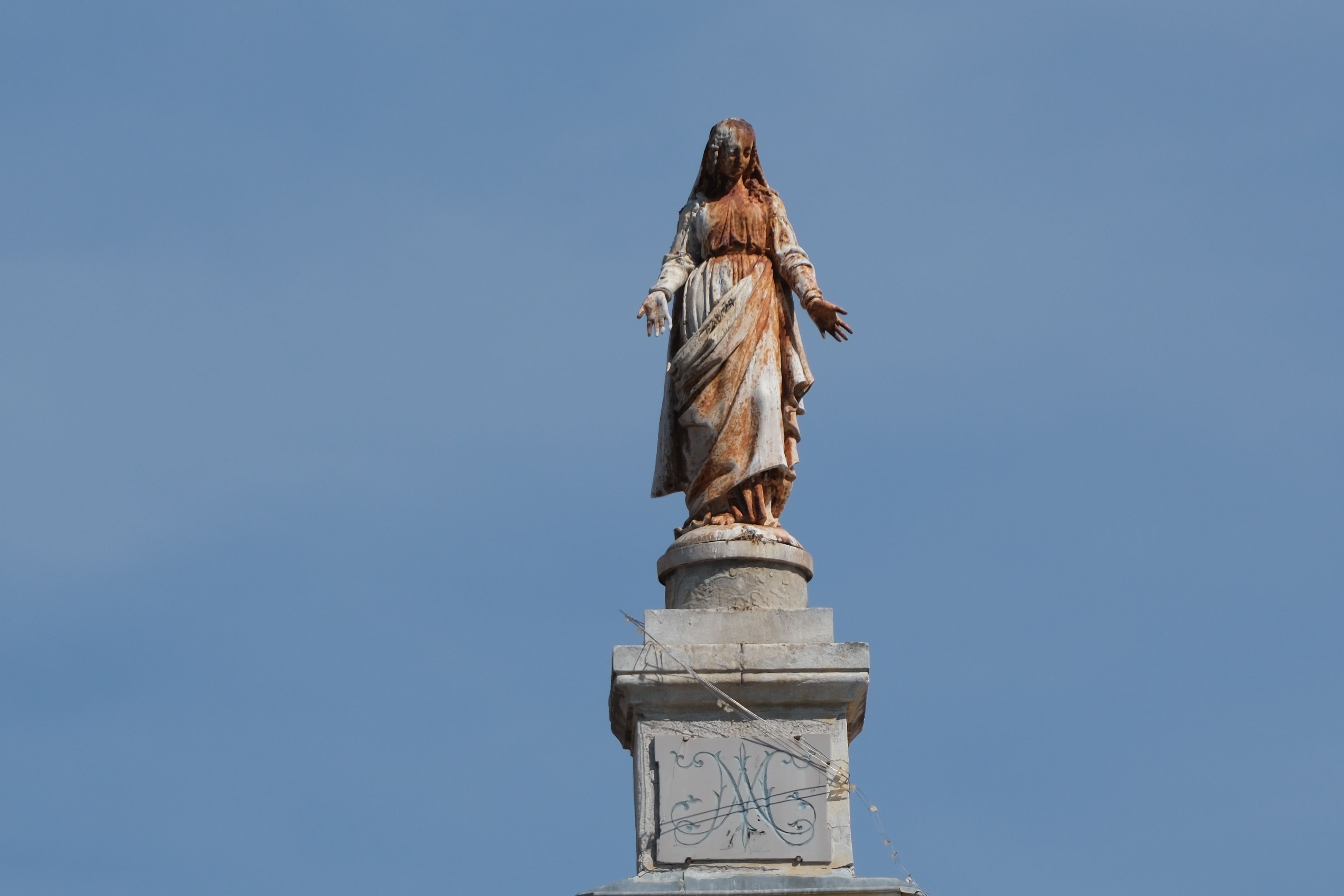 A weathered religious statue standing tall atop a column, set against a clear blue sky.