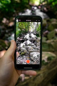 A hand holding a smartphone capturing a scenic waterfall in a lush green forest.