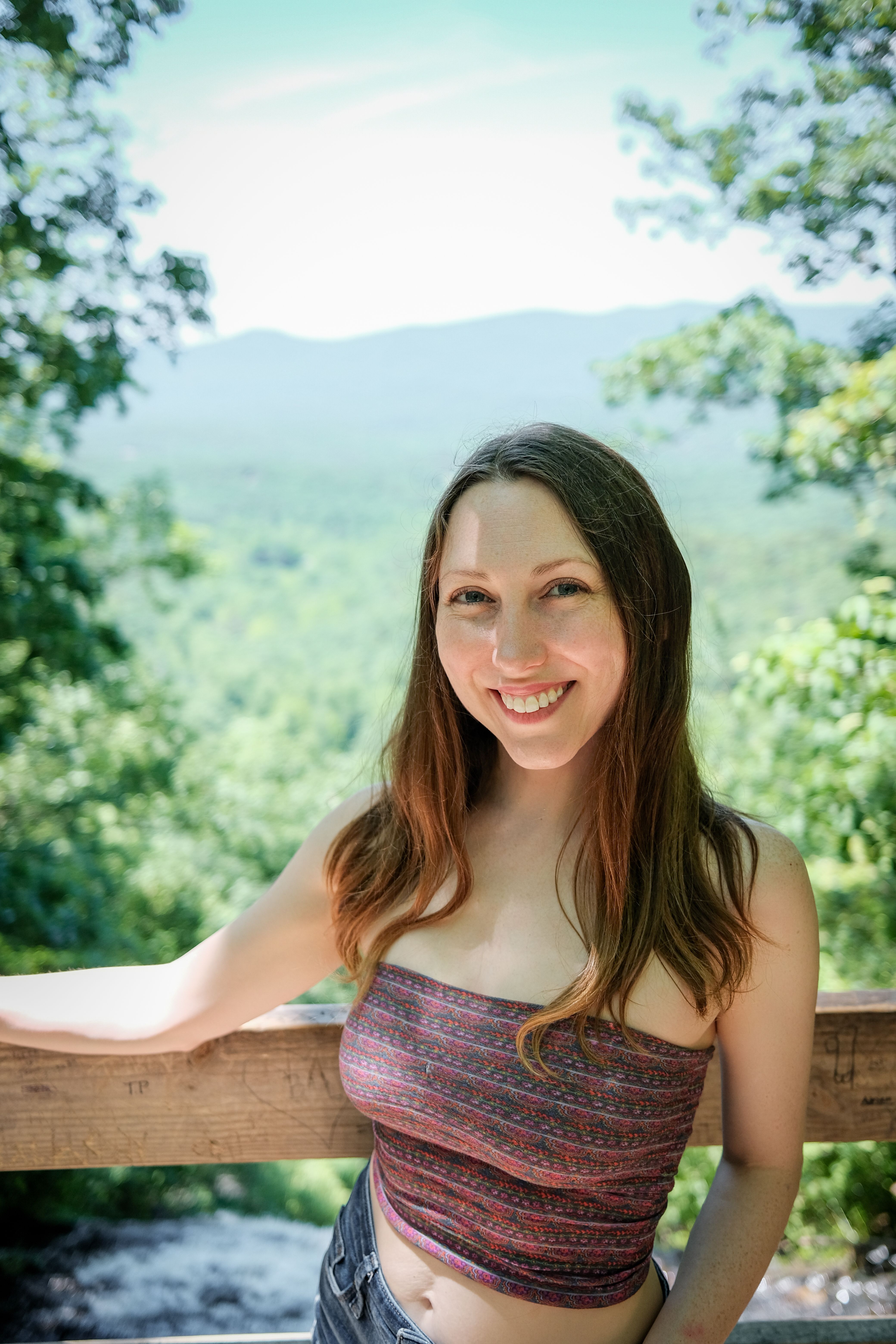 Smiling young woman posing against a lush mountainous landscape on a bright day.