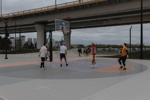 Men playing basketball on an urban court under a bridge on a cloudy day.