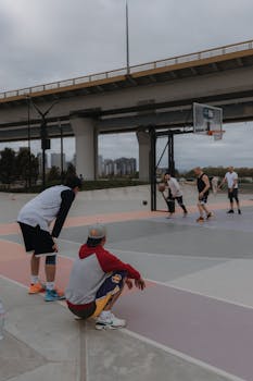 Group of adults playing basketball on an urban court under a bridge.