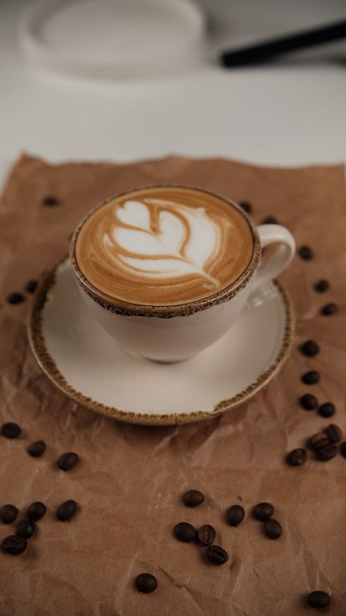 Free Close-up of a latte with froth art in a white cup surrounded by coffee beans. Stock Photo