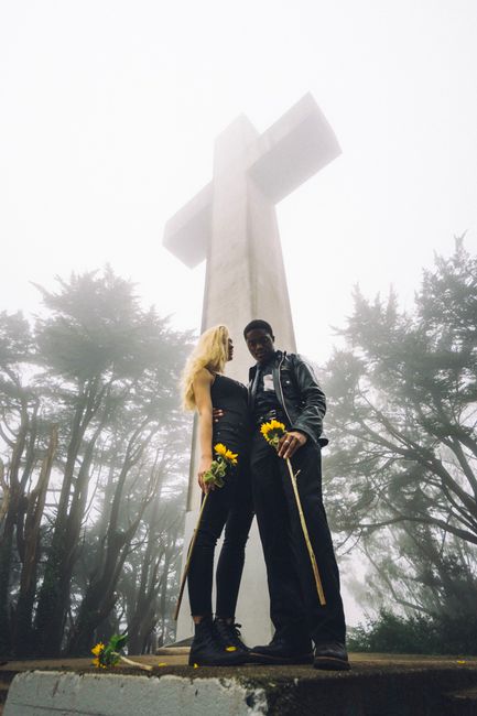 A couple holding sunflowers stands by a large cross in a foggy park, conveying romance.