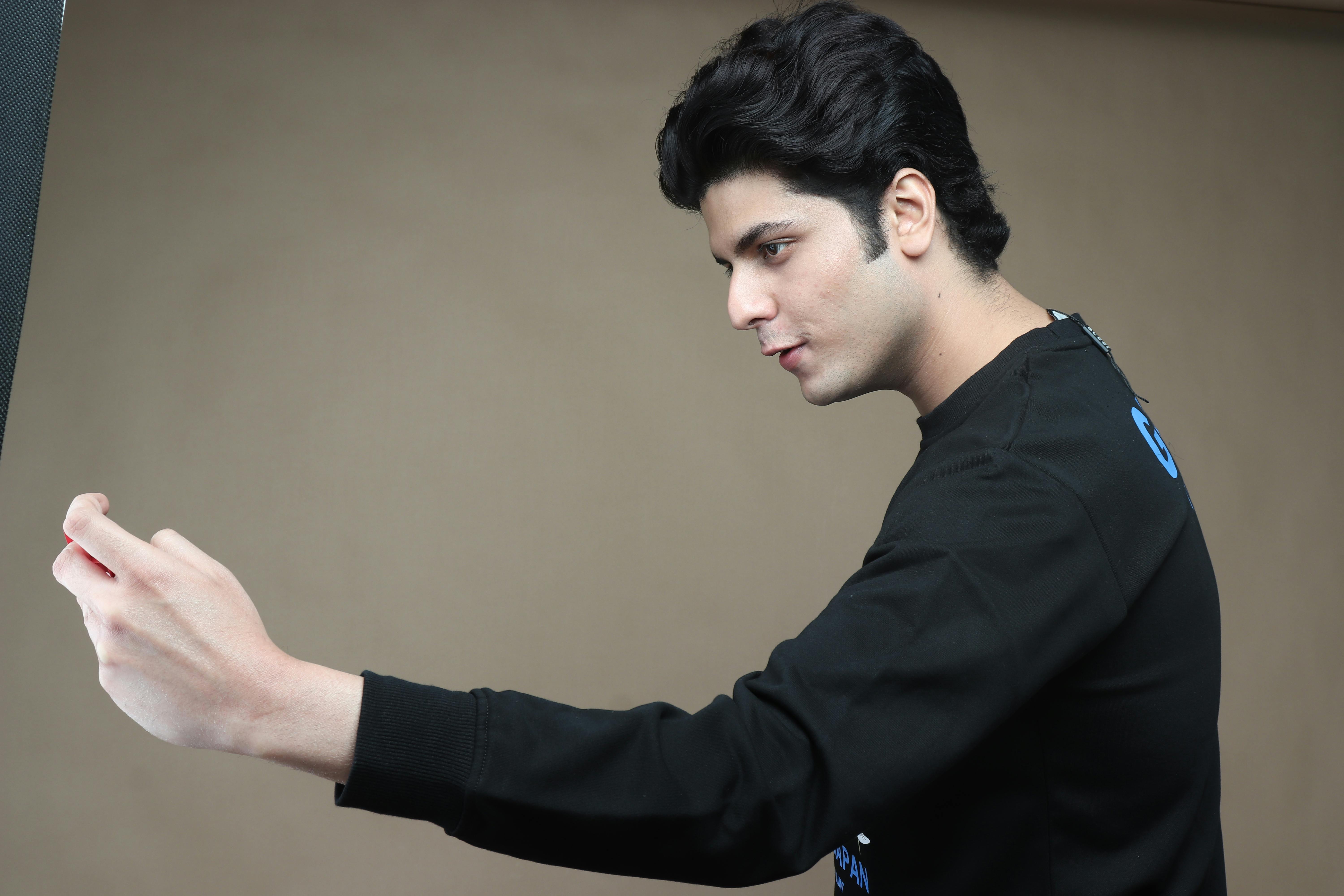Profile view of a young man with black hair taking a selfie indoors against a plain background.