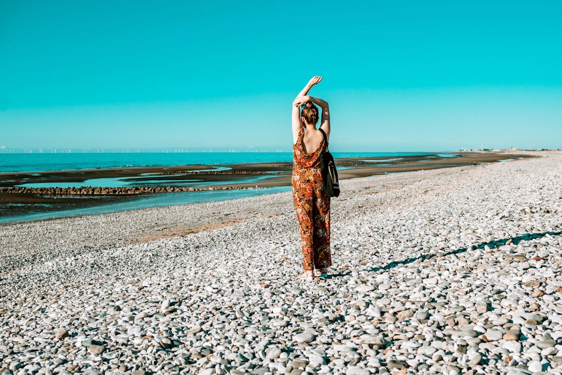 Free Back view photo of a woman wearing orange and black floral sleeveless jumpsuit standing on seashore Stock Photo