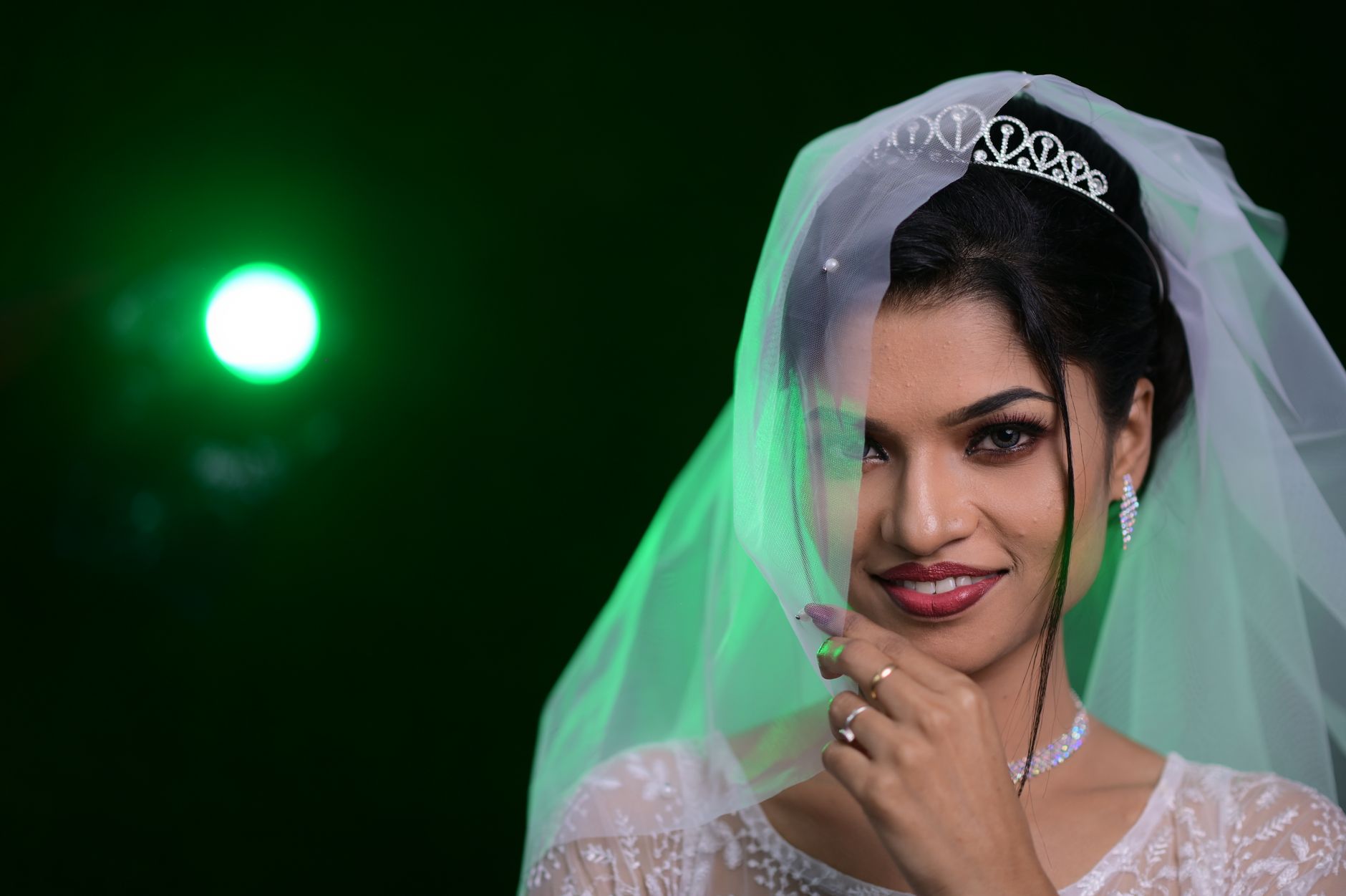 Close-up portrait of a smiling bride wearing a tiara and veil with a striking green light background.