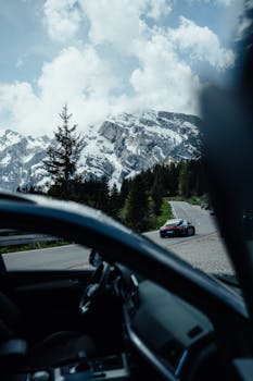 A car driving through a mountainous road with snow-capped peaks and evergreen trees under a clear blue sky.