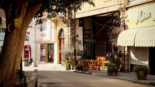 Free Sunlit city street with outdoor café seating and vibrant plants. Stock Photo