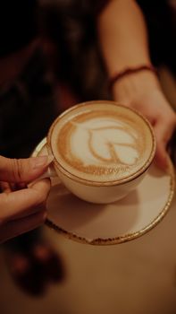 A woman holding a latte with intricate art in a porcelain cup and saucer.. woman, holding, latte