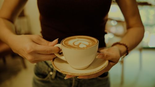 Free A close-up of hands holding a latte with heart-shaped art in a cozy setting. Stock Photo