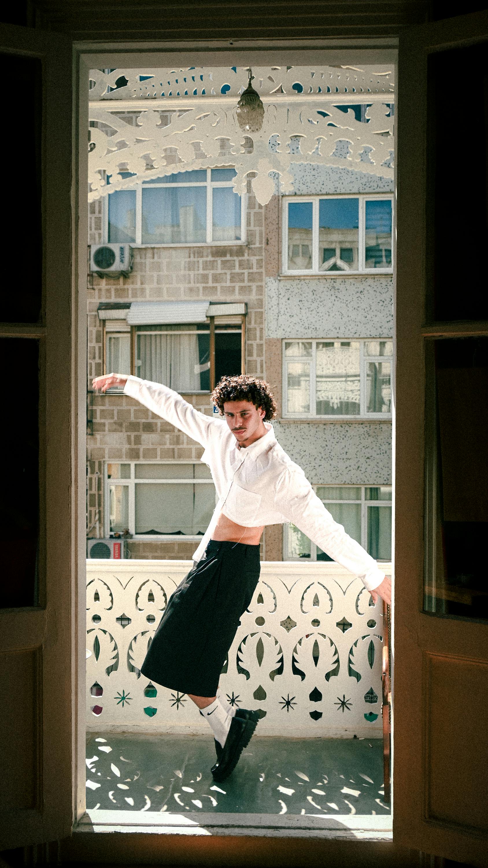 A fashionable man striking a pose on a sunlit balcony with urban backdrop.