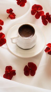 Aesthetic top view of a coffee cup with red flower petals on a white tablecloth.