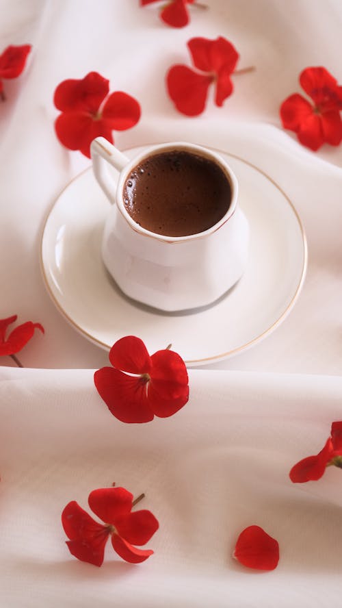 A cup of coffee on a white table with red petals