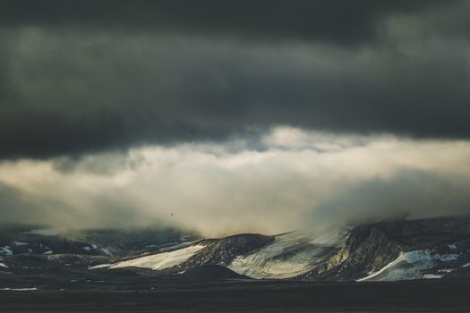 A moody landscape of mountains beneath a dramatic overcast sky full of thick clouds.